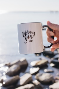 A person holds a white mug with the words 'Morning Vibes' written on it. The mug is positioned against a serene backdrop of water and rocks, reflecting a peaceful, early morning atmosphere.