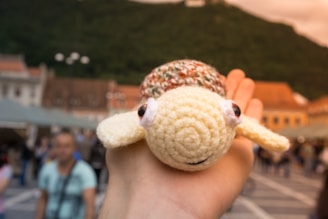 A hand holds a small crocheted turtle with a yellow body and brown shell against an outdoor backdrop of blurred people and buildings. A green hillside rises in the distance with a clear sky above.