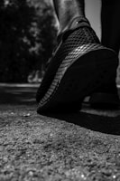 A close-up of a person's foot wearing a sports shoe, with an emphasis on the shoe's textured sole. The image is in black and white, highlighting the contrast between the footwear and the rough surface of the ground. Shadows and lighting create a dramatic effect, with trees and foliage blurred in the background.