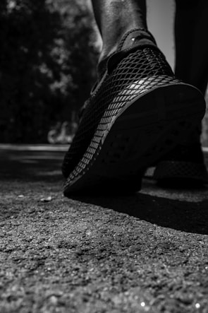 A close-up of a person's foot wearing a sports shoe, with an emphasis on the shoe's textured sole. The image is in black and white, highlighting the contrast between the footwear and the rough surface of the ground. Shadows and lighting create a dramatic effect, with trees and foliage blurred in the background.