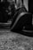 A close-up of a person's foot wearing a sports shoe, with an emphasis on the shoe's textured sole. The image is in black and white, highlighting the contrast between the footwear and the rough surface of the ground. Shadows and lighting create a dramatic effect, with trees and foliage blurred in the background.