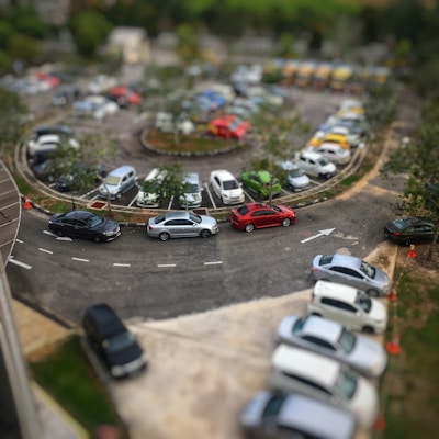 A circular parking lot is filled with a variety of cars arranged neatly in rows. Trees and small shrubs decorate the central area, adding greenery amidst the vehicles. A few traffic cones are visible, guiding the flow of cars. The scene is captured from an elevated viewpoint, providing a comprehensive view of the bustling parking area.