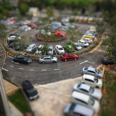 A circular parking lot is filled with a variety of cars arranged neatly in rows. Trees and small shrubs decorate the central area, adding greenery amidst the vehicles. A few traffic cones are visible, guiding the flow of cars. The scene is captured from an elevated viewpoint, providing a comprehensive view of the bustling parking area.