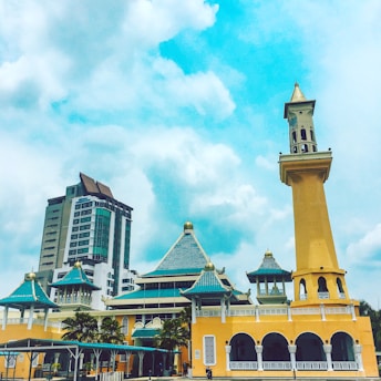 A large mosque with a modern architectural design featuring yellow walls, blue-green roofs topped with gold domes, and a tall minaret. In the background, there's a tall building that appears to be a modern office or residential structure. The sky is mostly clear with some scattered clouds, creating a bright and vibrant atmosphere. Palm trees and a small pavilion can be seen in front of the mosque.