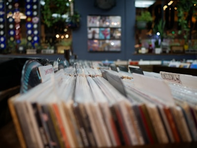 Rows of vinyl records are arranged in a record store, with labels like 'classical' visible. The background includes a blue wall, green plants, and blurred out decor and lighting.