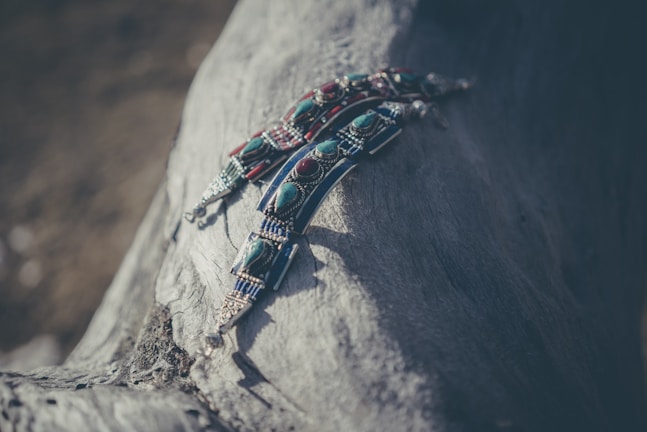 Intricately detailed beaded jewelry with turquoise and red stones laid on a textured piece of driftwood. The materials have a handcrafted, rustic feel, and the image is softly lit creating a tranquil and artistic atmosphere.