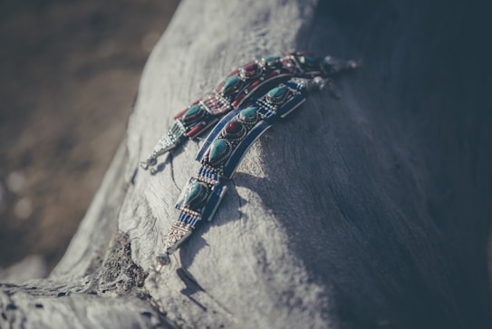 Intricately detailed beaded jewelry with turquoise and red stones laid on a textured piece of driftwood. The materials have a handcrafted, rustic feel, and the image is softly lit creating a tranquil and artistic atmosphere.