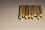 Eight colored pencils with natural wood casing are neatly aligned on a textured, pale surface. Each pencil displays a different color at the tip, including black, red, green, and yellow.