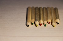 Eight colored pencils with natural wood casing are neatly aligned on a textured, pale surface. Each pencil displays a different color at the tip, including black, red, green, and yellow.