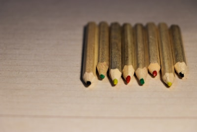 Eight colored pencils with natural wood casing are neatly aligned on a textured, pale surface. Each pencil displays a different color at the tip, including black, red, green, and yellow.