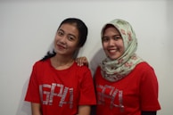 Two smiling individuals are standing against a plain white background. They are both wearing red shirts with printed texts and one of them is wearing a patterned hijab.
