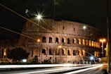 A historic amphitheater stands illuminated against the night sky, with bright streetlights casting a warm glow across its ancient arches. Light streaks from passing vehicles create dynamic lines along the road in the foreground.