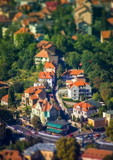 A picturesque aerial view of a small urban neighborhood featuring charming houses with orange-tiled roofs nestled among lush greenery. The vibrant scene captures a winding street flanked by well-maintained homes and leafy trees, creating a serene and idyllic atmosphere.