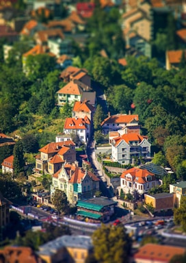 A picturesque aerial view of a small urban neighborhood featuring charming houses with orange-tiled roofs nestled among lush greenery. The vibrant scene captures a winding street flanked by well-maintained homes and leafy trees, creating a serene and idyllic atmosphere.