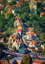 A picturesque aerial view of a small urban neighborhood featuring charming houses with orange-tiled roofs nestled among lush greenery. The vibrant scene captures a winding street flanked by well-maintained homes and leafy trees, creating a serene and idyllic atmosphere.