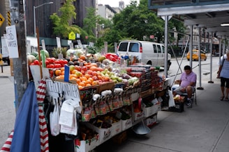 A street vendor stand filled with a variety of fresh fruits and vegetables, including tomatoes, oranges, apples, and pineapples. Baskets of produce are neatly arranged on a table covered with white and brown boxes. Signs displaying prices are visible, and there are signs advertising ice-cold water. A man in a pink shirt is seated on a folding chair next to the stand under some scaffolding, while pedestrians walk by. A white van and a yellow taxi are on the street, with trees and urban buildings in the background.