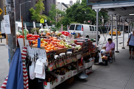 A street vendor stand filled with a variety of fresh fruits and vegetables, including tomatoes, oranges, apples, and pineapples. Baskets of produce are neatly arranged on a table covered with white and brown boxes. Signs displaying prices are visible, and there are signs advertising ice-cold water. A man in a pink shirt is seated on a folding chair next to the stand under some scaffolding, while pedestrians walk by. A white van and a yellow taxi are on the street, with trees and urban buildings in the background.