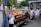 A street vendor stand filled with a variety of fresh fruits and vegetables, including tomatoes, oranges, apples, and pineapples. Baskets of produce are neatly arranged on a table covered with white and brown boxes. Signs displaying prices are visible, and there are signs advertising ice-cold water. A man in a pink shirt is seated on a folding chair next to the stand under some scaffolding, while pedestrians walk by. A white van and a yellow taxi are on the street, with trees and urban buildings in the background.