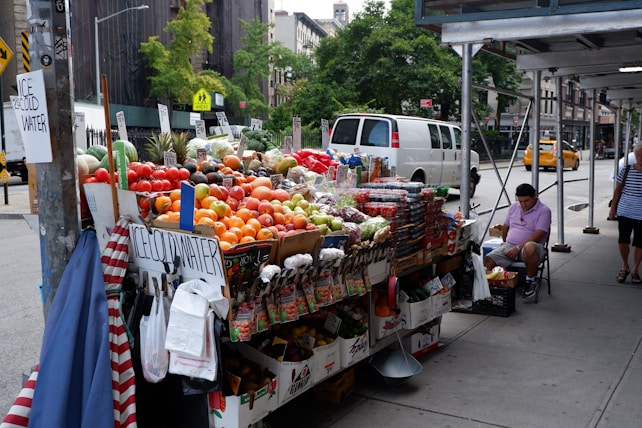 A street vendor stand filled with a variety of fresh fruits and vegetables, including tomatoes, oranges, apples, and pineapples. Baskets of produce are neatly arranged on a table covered with white and brown boxes. Signs displaying prices are visible, and there are signs advertising ice-cold water. A man in a pink shirt is seated on a folding chair next to the stand under some scaffolding, while pedestrians walk by. A white van and a yellow taxi are on the street, with trees and urban buildings in the background.