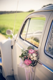 A vintage white car adorned with a floral arrangement on the door handle. The bouquet includes pink and white roses, green leaves, and small pink berries tied with a purple ribbon. The background features a blurred landscape with green fields under a sunny sky.