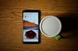 A smartphone displaying a gallery with images and a white mug with a green handle sitting on a wooden surface. The phone screen shows photos with captions and likes information.