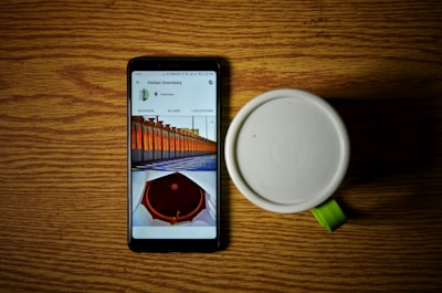 A smartphone displaying a gallery with images and a white mug with a green handle sitting on a wooden surface. The phone screen shows photos with captions and likes information.