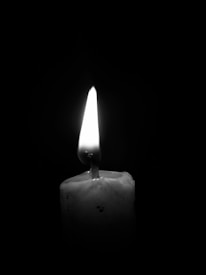 A single lit candle stands against a dark background, casting a soft glow. The flame is bright and flickers gently, causing shadows to dance on the wax surface. The wax appears to have some drips and imperfections, adding texture to the image.