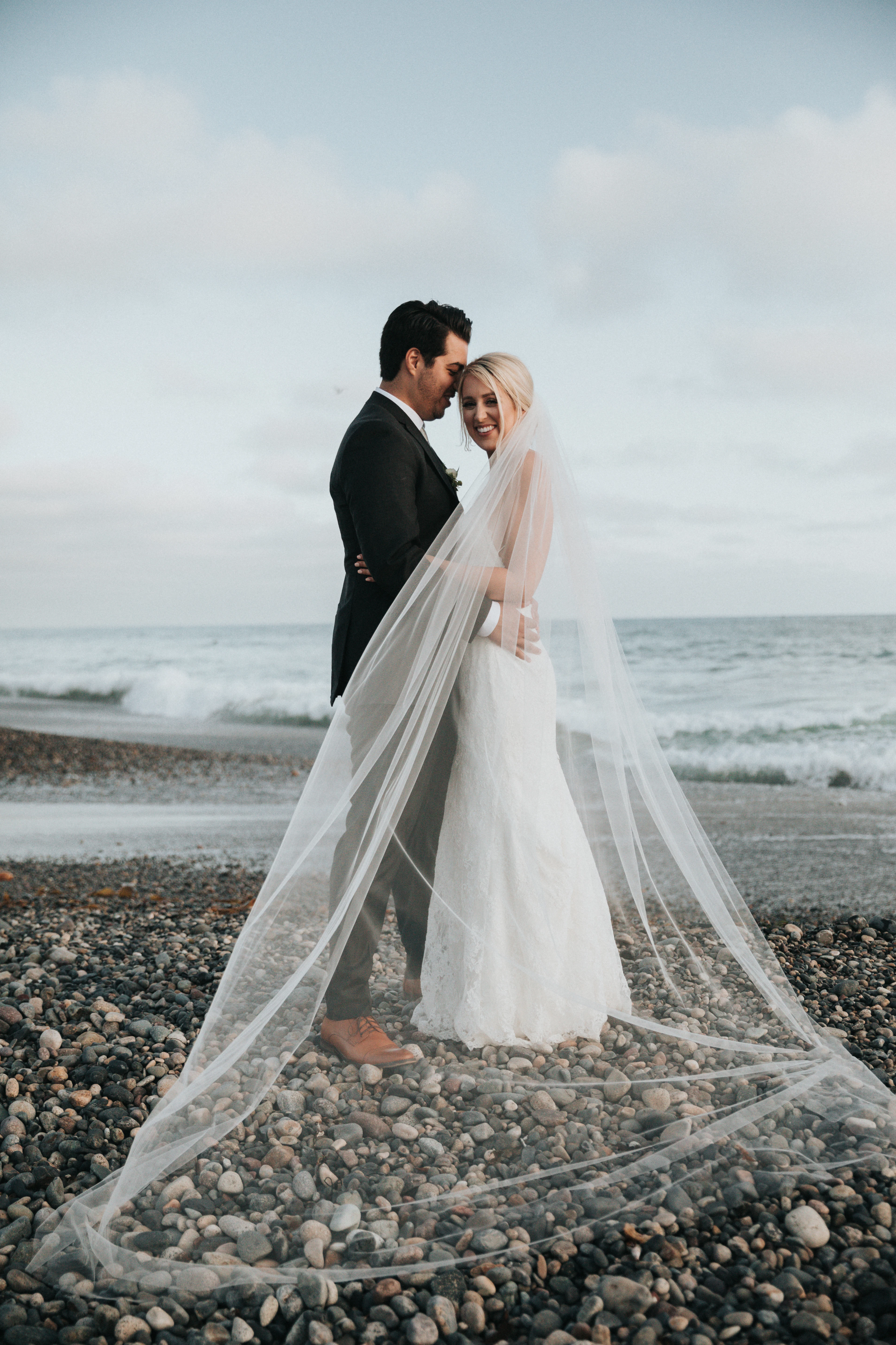 A couple at the water's edge, the bride's veil trailing behind her