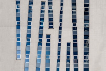 Tall narrow windows with blue-tinted glass are set vertically in an abstract pattern on a modern concrete building facade. The windows are of varying heights and are placed at slight angles, creating a dynamic visual effect.