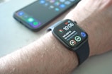 A smartwatch is worn on a wrist, displaying the time, date, and various health-related data points on its screen. Behind it, a smartphone with a colorful display sits on a surface.