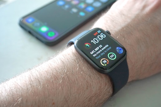 A smartwatch is worn on a wrist, displaying the time, date, and various health-related data points on its screen. Behind it, a smartphone with a colorful display sits on a surface.