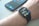 A smartwatch is worn on a wrist, displaying the time, date, and various health-related data points on its screen. Behind it, a smartphone with a colorful display sits on a surface.
