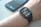 A smartwatch is worn on a wrist, displaying the time, date, and various health-related data points on its screen. Behind it, a smartphone with a colorful display sits on a surface.