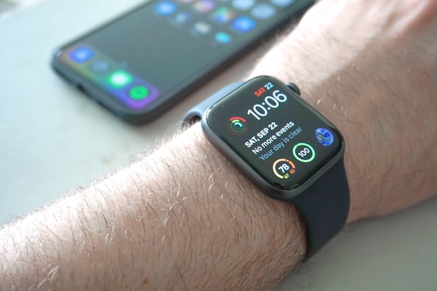A smartwatch is worn on a wrist, displaying the time, date, and various health-related data points on its screen. Behind it, a smartphone with a colorful display sits on a surface.
