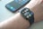 A smartwatch is worn on a wrist, displaying the time, date, and various health-related data points on its screen. Behind it, a smartphone with a colorful display sits on a surface.