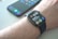 A smartwatch is worn on a wrist, displaying the time, date, and various health-related data points on its screen. Behind it, a smartphone with a colorful display sits on a surface.