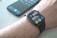 A smartwatch is worn on a wrist, displaying the time, date, and various health-related data points on its screen. Behind it, a smartphone with a colorful display sits on a surface.