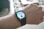 A close-up view of a person's wrist wearing a smartwatch with a black strap. The watch face displays various digital information, including a circular dial showing time, a fitness activity ring, and temperature readings. The background is blurred but hints at an indoor setting with a patterned rug and furniture.