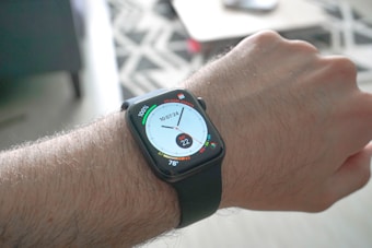 A close-up view of a person's wrist wearing a smartwatch with a black strap. The watch face displays various digital information, including a circular dial showing time, a fitness activity ring, and temperature readings. The background is blurred but hints at an indoor setting with a patterned rug and furniture.