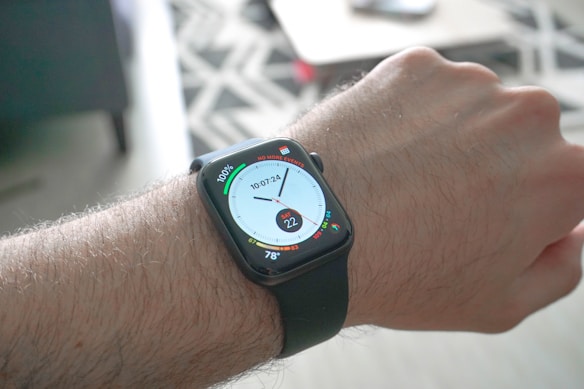 A close-up view of a person's wrist wearing a smartwatch with a black strap. The watch face displays various digital information, including a circular dial showing time, a fitness activity ring, and temperature readings. The background is blurred but hints at an indoor setting with a patterned rug and furniture.