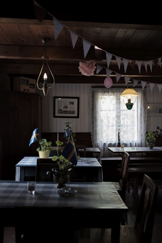 An interior of a rustic room with wooden beams and a plaid tablecloth on the tables. Small potted plants with miniature Swedish flags decorate the tables. A garland of triangular flags and a single pink balloon hang from the ceiling. Two pendant lights provide dim illumination, enhancing the cozy atmosphere. A window with sheer curtains allows diffused natural light to enter the room.