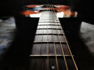 A close-up perspective of a guitar, focusing on the neck and the strings. The lighting creates a reflective sheen on the wood surface, with the guitar body partially visible. The background is slightly blurred, emphasizing the instrument’s details.