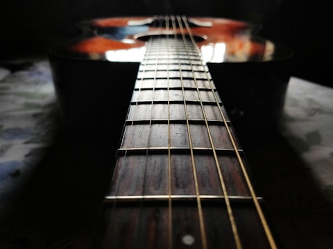 A close-up perspective of a guitar, focusing on the neck and the strings. The lighting creates a reflective sheen on the wood surface, with the guitar body partially visible. The background is slightly blurred, emphasizing the instrument’s details.