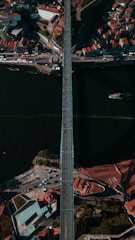 Aerial view of a long bridge spanning across a wide, dark river with a dense urban area on either side. Buildings with red-tiled roofs cluster around the bridge's approaches, and there are small boats visible on the river. The scene is bustling with activity including cars and people on the bridge.