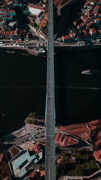 Aerial view of a long bridge spanning across a wide, dark river with a dense urban area on either side. Buildings with red-tiled roofs cluster around the bridge's approaches, and there are small boats visible on the river. The scene is bustling with activity including cars and people on the bridge.