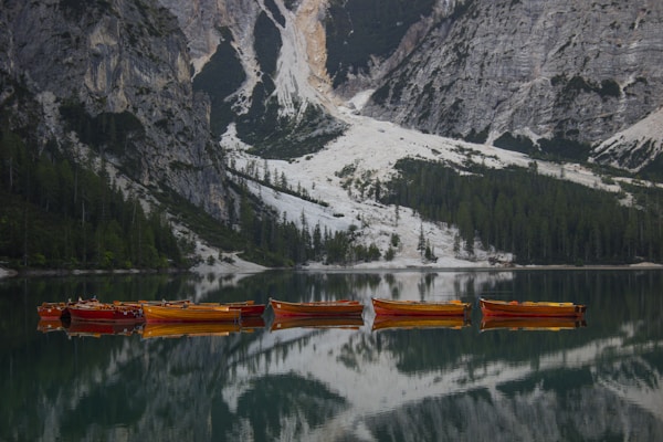 A tranquil lake with several wooden boats floating in a line. The water is calm, reflecting the steep surrounding cliffs and dense evergreen forests. The mountains have rugged rock faces and patches of greenery.