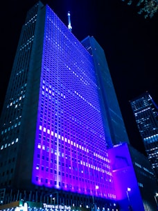 A tall skyscraper is illuminated with vibrant blue and purple lights against a dark night sky. Numerous windows are visible, forming a grid pattern across the building's facade. The top of the building features a lit spire. In the foreground, there is signage indicating a location called Tavern at the Park.