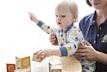 A young child wearing a patterned onesie is interacting with colorful alphabet blocks on a table. An adult sitting beside the child is supporting them, dressed in a navy blue shirt with an embroidered logo. The scene conveys a sense of learning or playtime in a bright, softly-lit room.