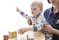 A young child wearing a patterned onesie is interacting with colorful alphabet blocks on a table. An adult sitting beside the child is supporting them, dressed in a navy blue shirt with an embroidered logo. The scene conveys a sense of learning or playtime in a bright, softly-lit room.