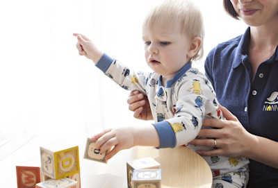 A young child wearing a patterned onesie is interacting with colorful alphabet blocks on a table. An adult sitting beside the child is supporting them, dressed in a navy blue shirt with an embroidered logo. The scene conveys a sense of learning or playtime in a bright, softly-lit room.